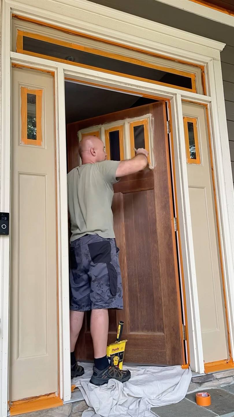 A worker refinishing a timber front door in its frame, the threshold masked and a drop cloth down