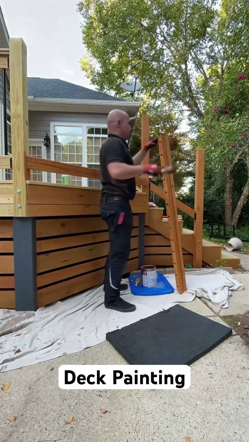 A worker staining the posts and horizontal skirt boards of a newly built deck, a drop cloth over the path