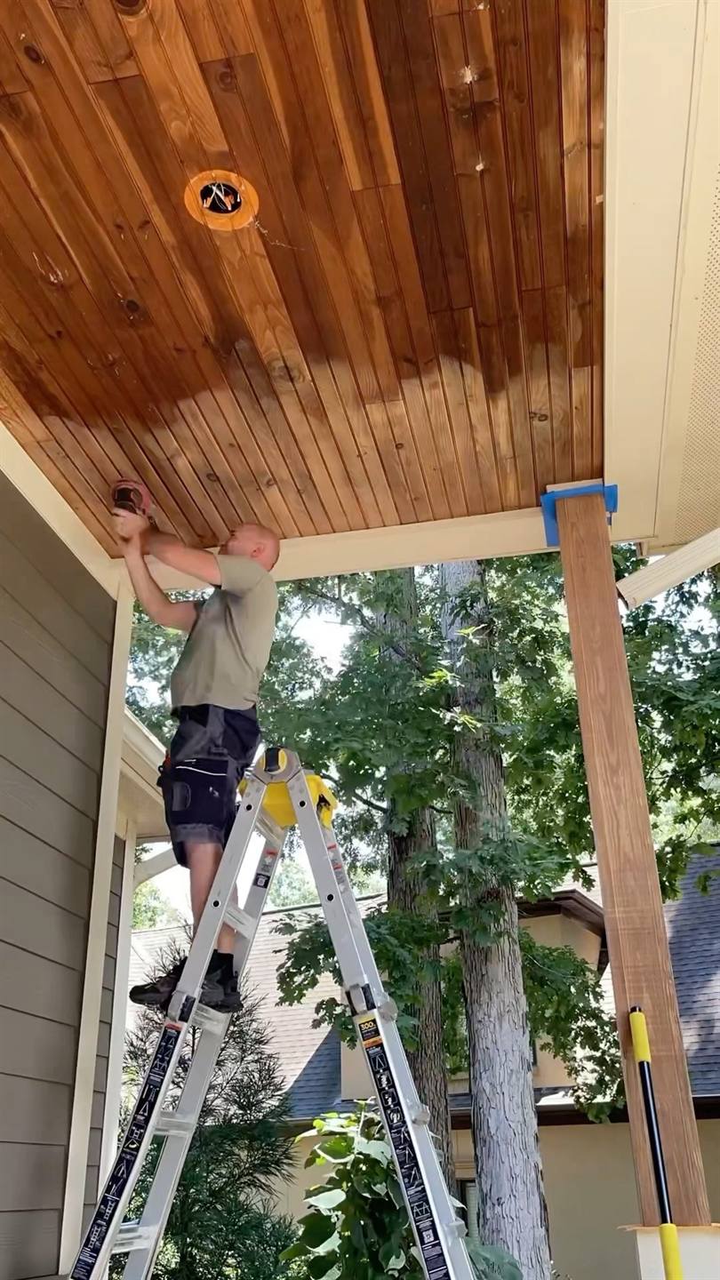 A worker on a ladder sanding the stained boards of a porch ceiling back to bare timber