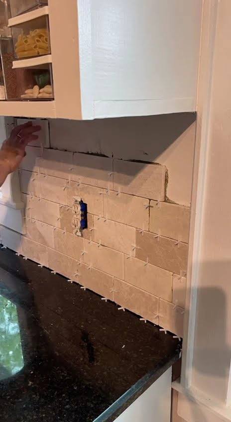 Backsplash tile being pressed onto the wall above a dark granite counter beside a pale cabinet run