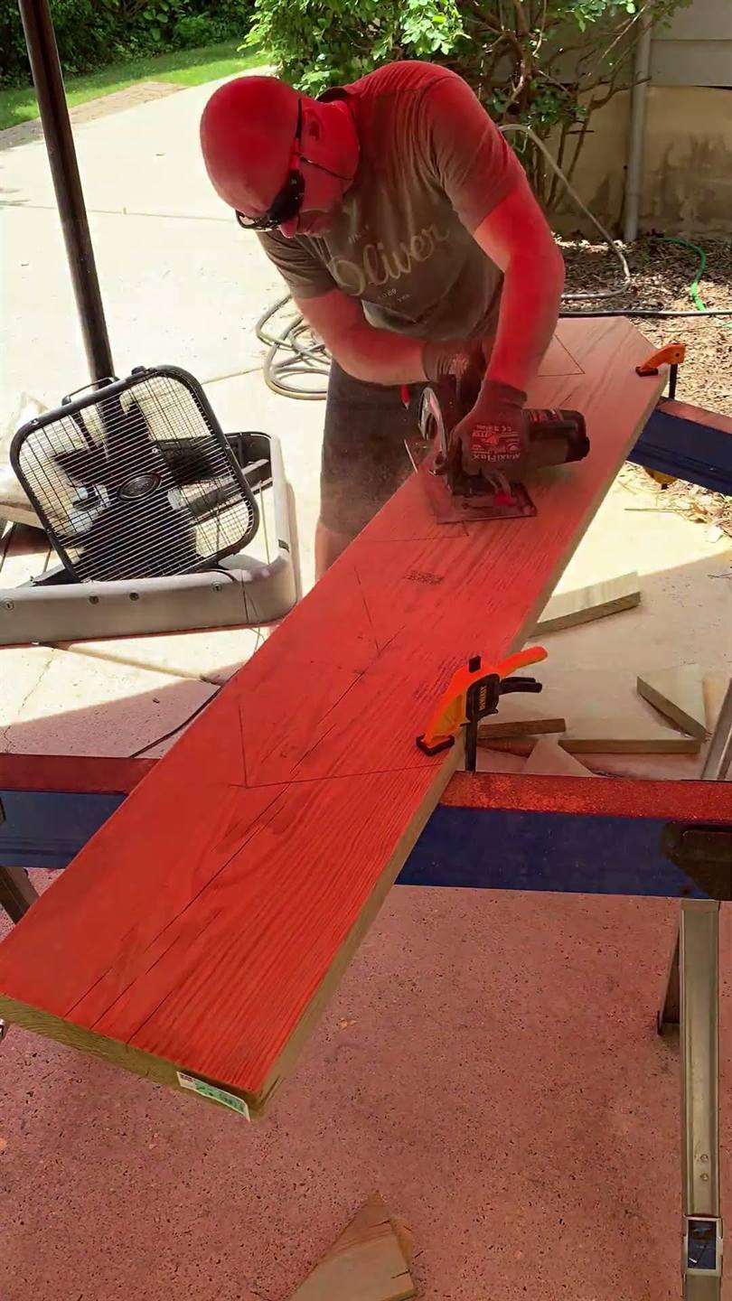 A carpenter cutting a long red-stained board to length with a circular saw on a work stand in a driveway