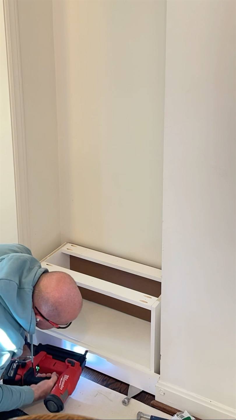 A carpenter kneeling to fix the white base frame of a built-in cabinet to the floor with a nail gun