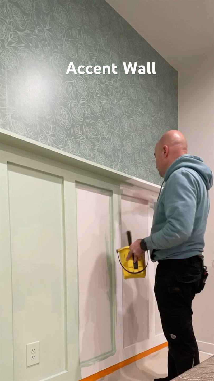 A painter cutting in pale green paint on framed wall paneling below a patterned wallpaper above the rail