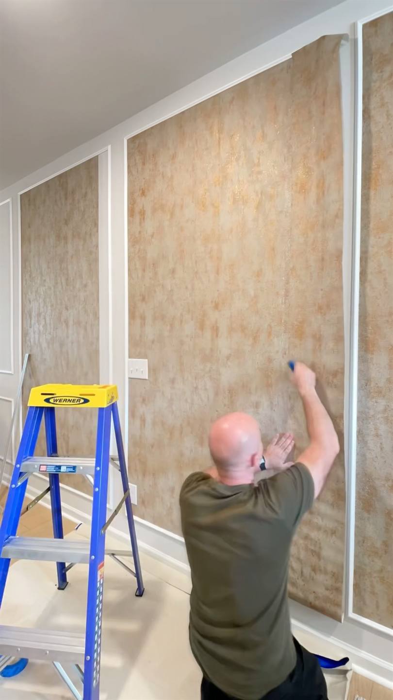 A worker on a stepladder pressing a wood-grain wall panel into place between white trim in a hallway