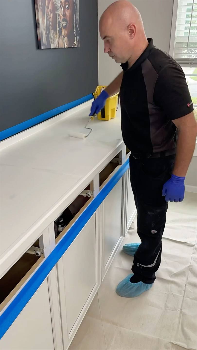 A worker in gloves masking a kitchen counter with blue tape, the cabinets below covered and the floor sheeted