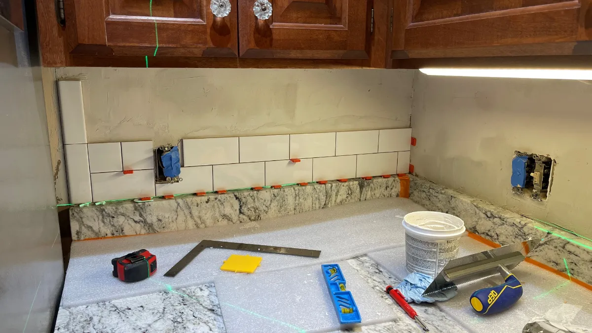The first two courses of cream subway tile set to a laser line, with spacers, a square and a level on the papered counter