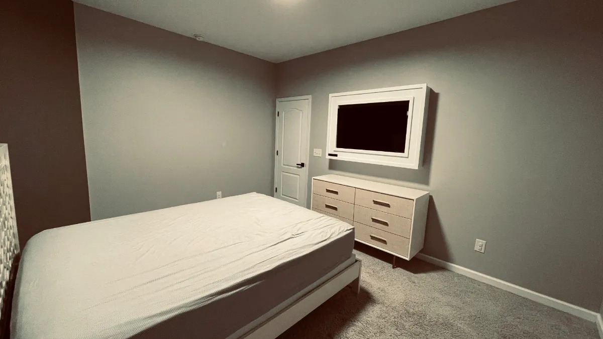 A bedroom in deep brown with a made bed and a pale dresser under a mounted screen